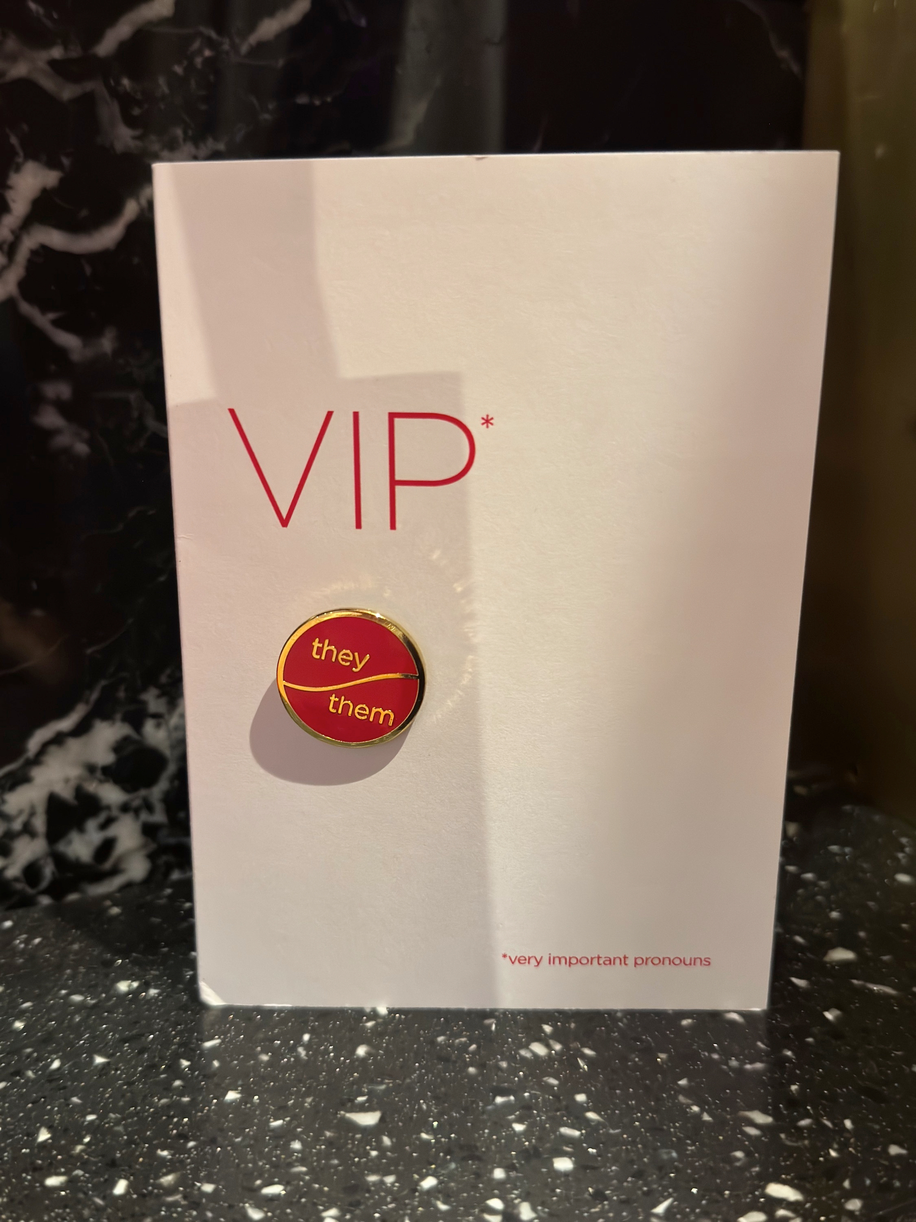 A white card reading “VIP*” with a red enamel pin labeled “they them,” resting on a dark stone surface, with small text noting “very important pronouns.”