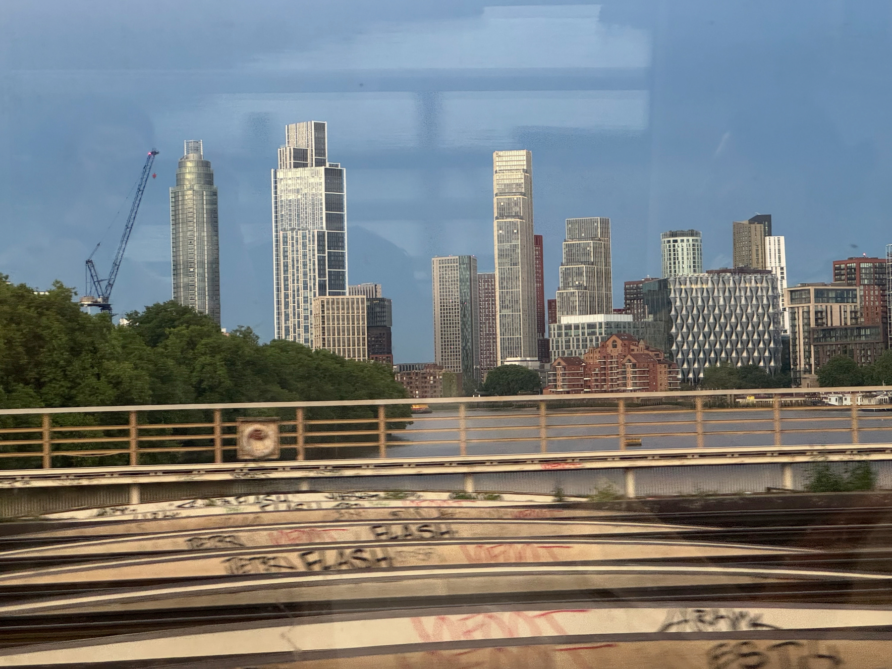 City skyline seen through a train window, with tall modern towers clustered near the river, cranes in the distance, and blurred tracks and railings in the foreground suggesting motion.