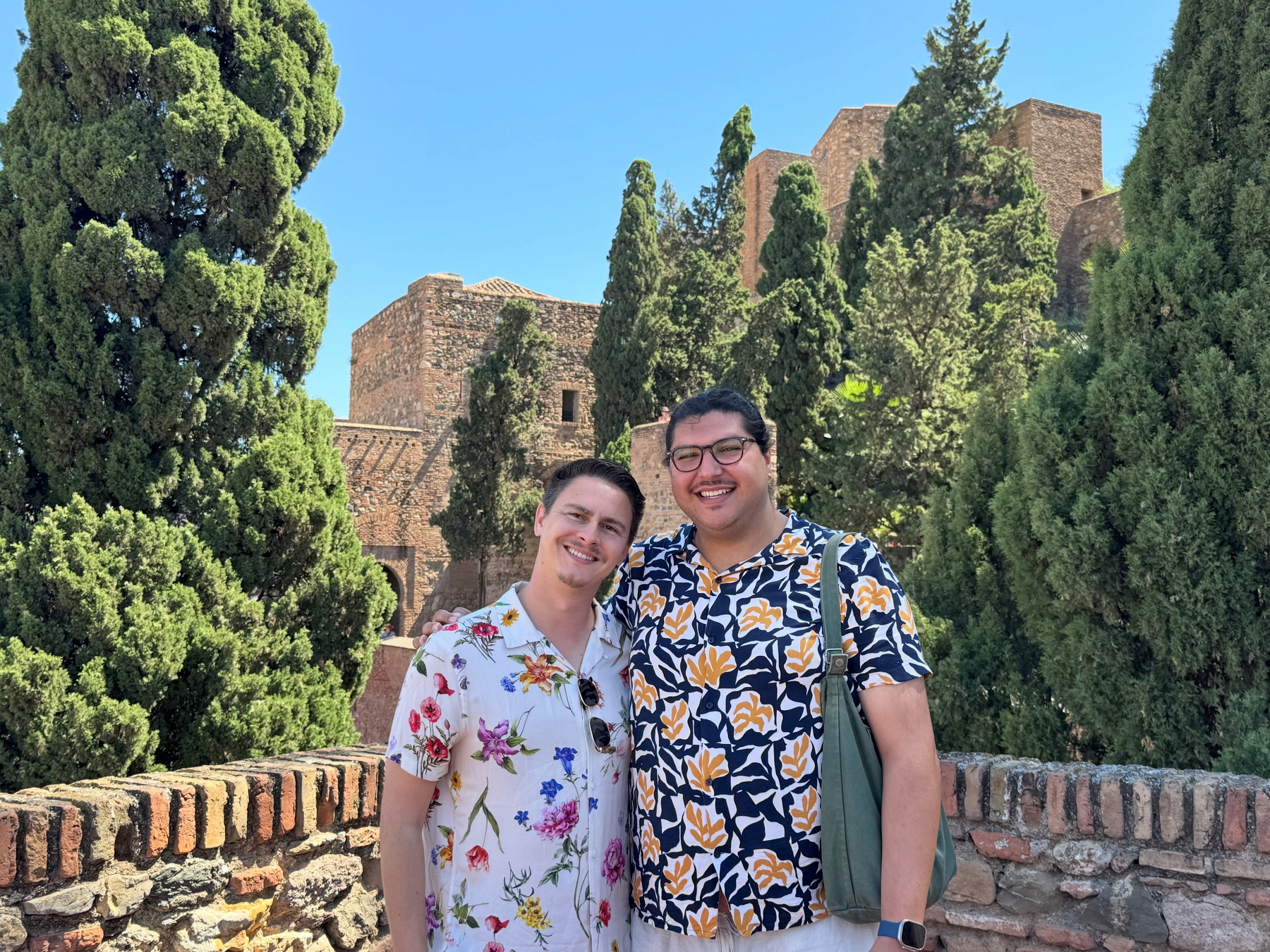 Hugo and I standing arm in arm on a stone terrace surrounded by tall trees, with historic fortress walls and towers rising behind us in bright daylight.