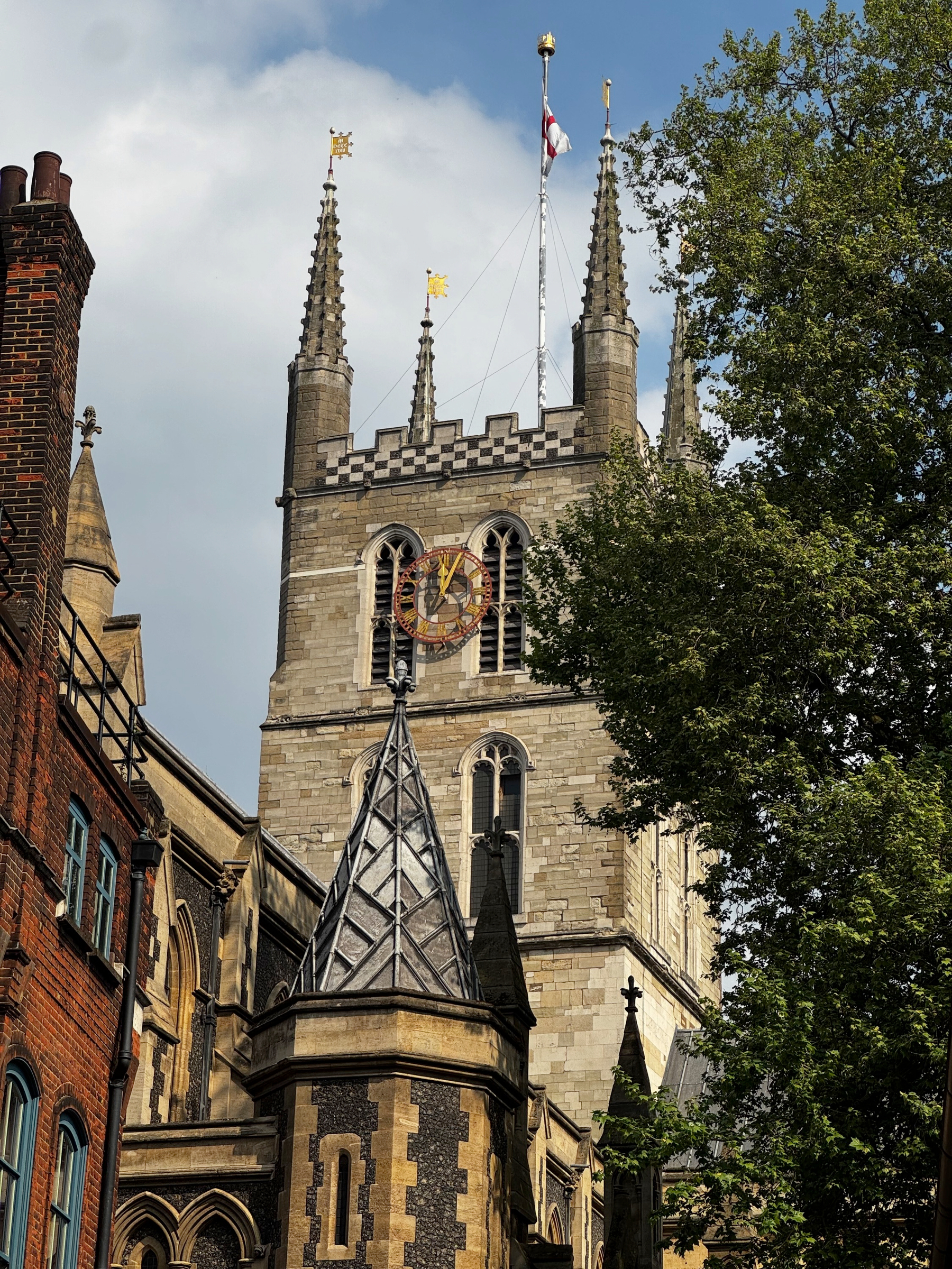 Historic church tower with a large clock face and four stone pinnacles, partially framed by trees, rising above surrounding buildings under a soft afternoon sky.