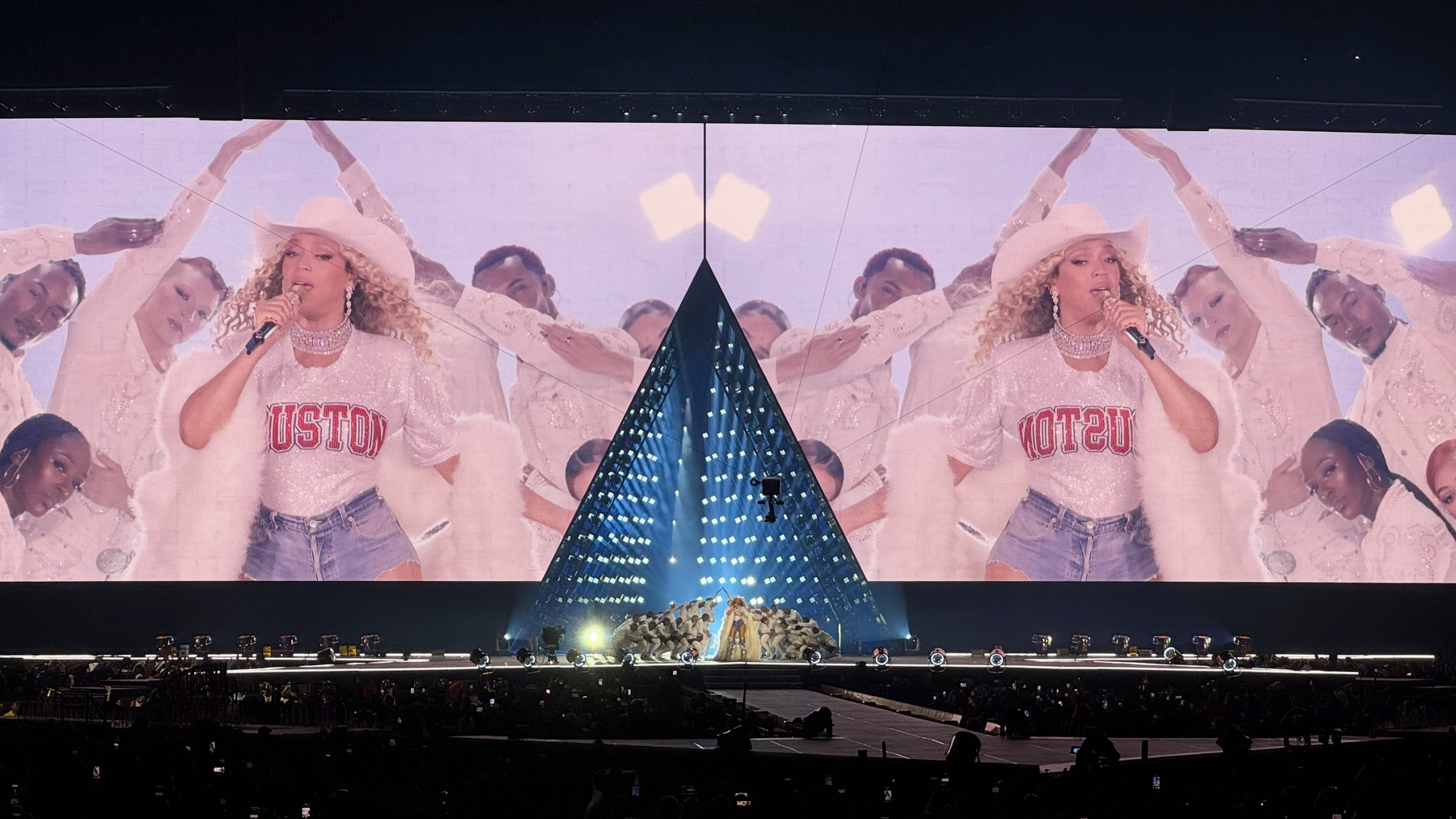 Beyoncé performs on a massive stadium stage with two enormous mirrored screens showing her in a white cowboy hat, sparkly Houston jersey-style top, denim shorts, and feathered coat, singing into a microphone while dancers frame her. A glowing triangular stage structure anchors the center, with dramatic lighting and a sea of fans holding up phones in the darkened stadium.