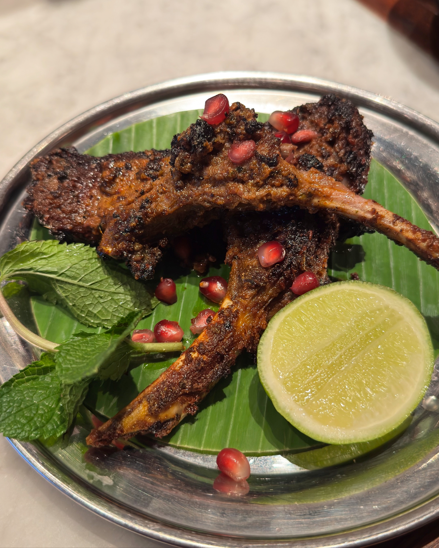 Tandoori Lamb Chops with deeply caramelized, charred edges are arranged on a banana leaf, topped with ruby-red pomegranate seeds and served with a slice of lime and fresh mint. The dark crust and vibrant garnishes emphasize the meat’s richness and smoky, aromatic appeal.
