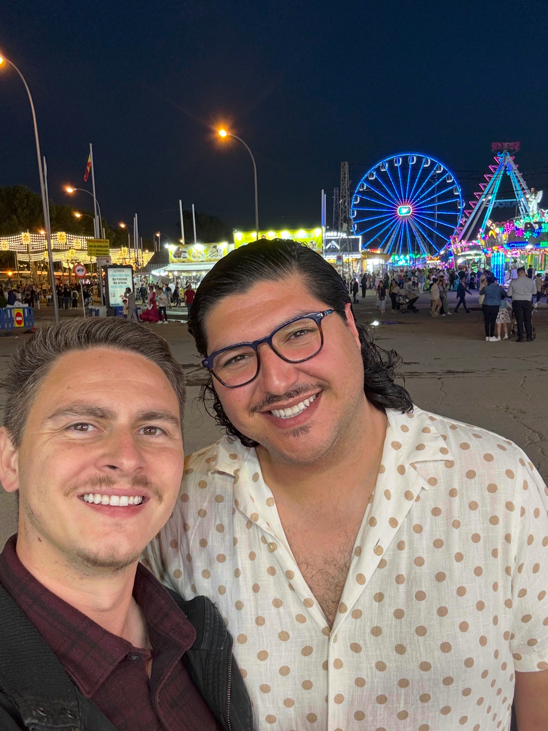 Hugo and I smiling at night with a brightly lit fair behind us, featuring a glowing Ferris wheel, colorful rides, and crowds moving through the illuminated grounds.