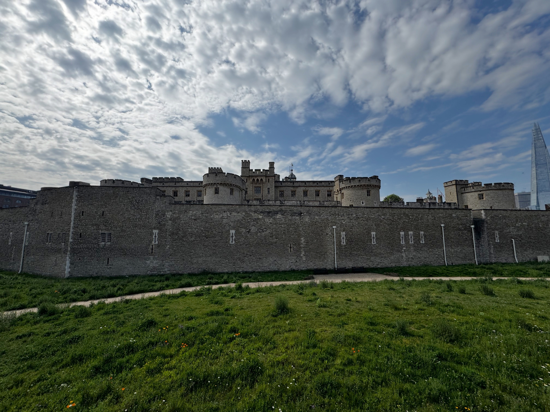 A long stretch of weathered stone curtain wall and round towers fills the frame beneath dramatic, textured clouds, while the glassy peak of the Shard appears in the distance, emphasizing the contrast between historic fortifications and modern London.