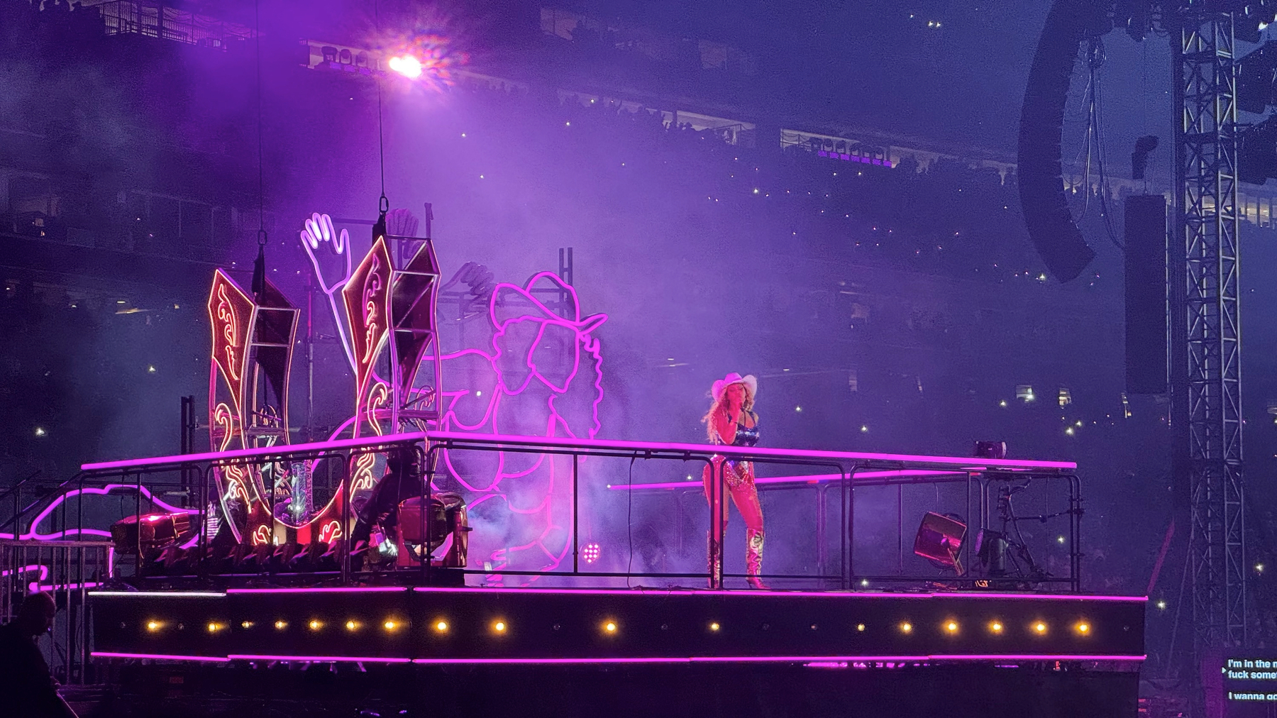 Beyoncé stands confidently on an extended runway platform, wearing a cowboy hat and sparkling outfit, surrounded by purple haze and neon Western-style graphics, including a large horseshoe motif. The elevated platform lights and mist create a dreamy, cinematic Western atmosphere.