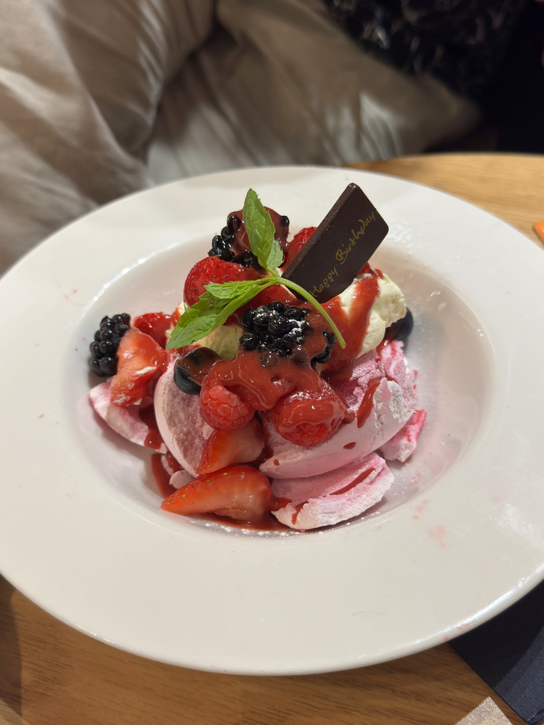 A generous berry dessert layered with pink meringue, cream, and fresh strawberries, raspberries, and blackberries, topped with mint and a chocolate plaque, lightly dusted with powdered sugar.