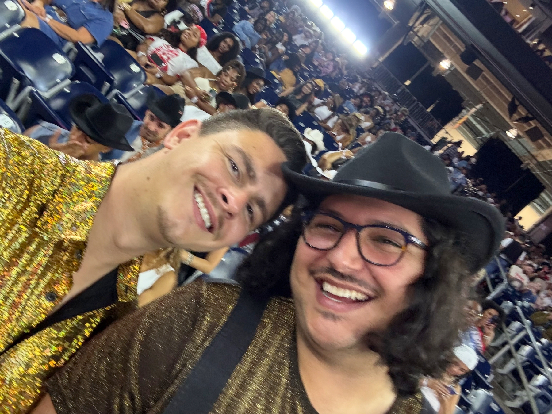 Two smiling concertgoers take a close-up selfie in the stadium stands, wearing glittery gold outfits and cowboy hats, with rows of excited fans visible behind them. The image captures the joyful anticipation and communal energy of the crowd before or during the show.