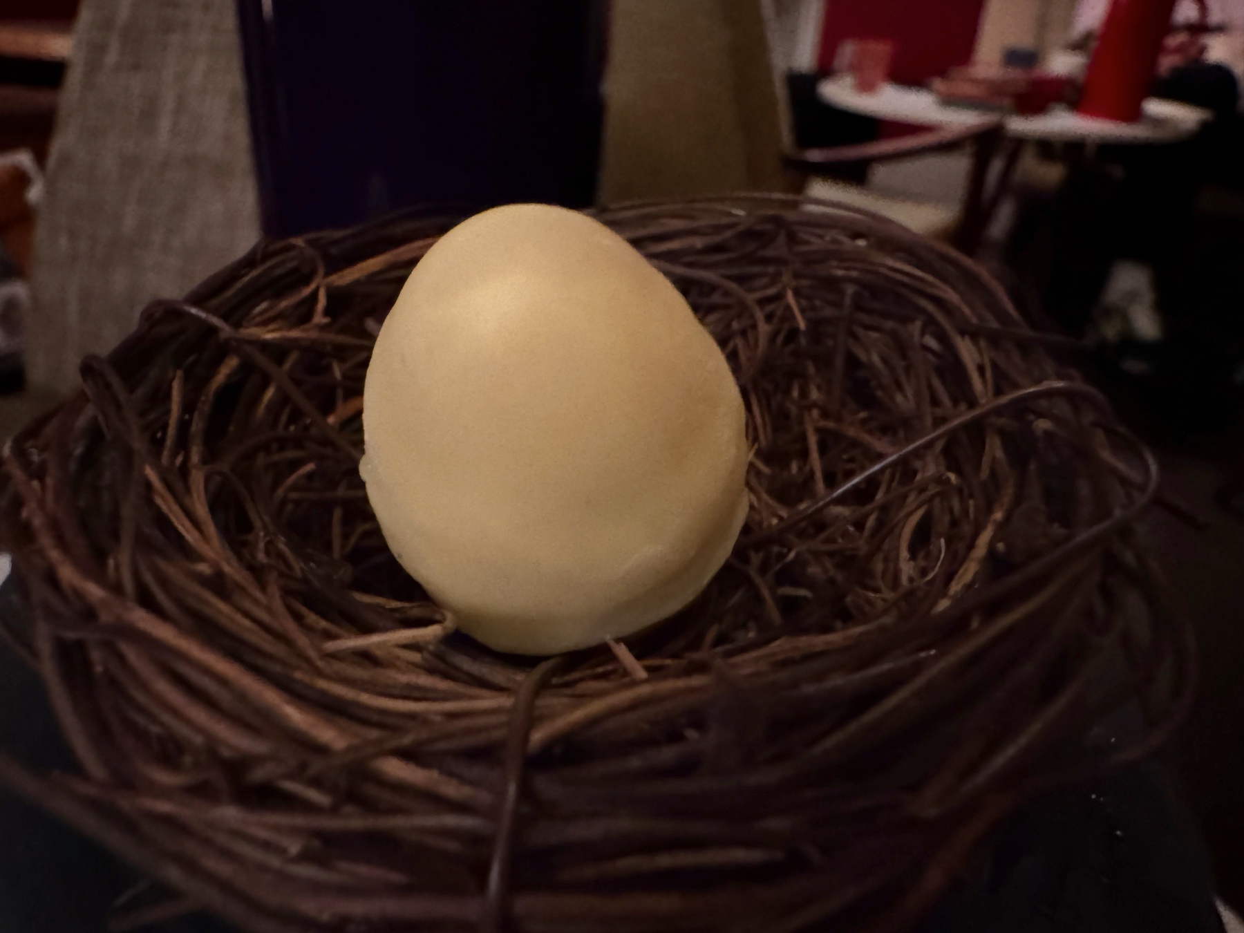 A smooth, pale yellow clarified cocktail sphere resting in a tightly woven twig nest, its matte surface and careful placement giving it the feel of an edible sculpture served as part of a tasting course.