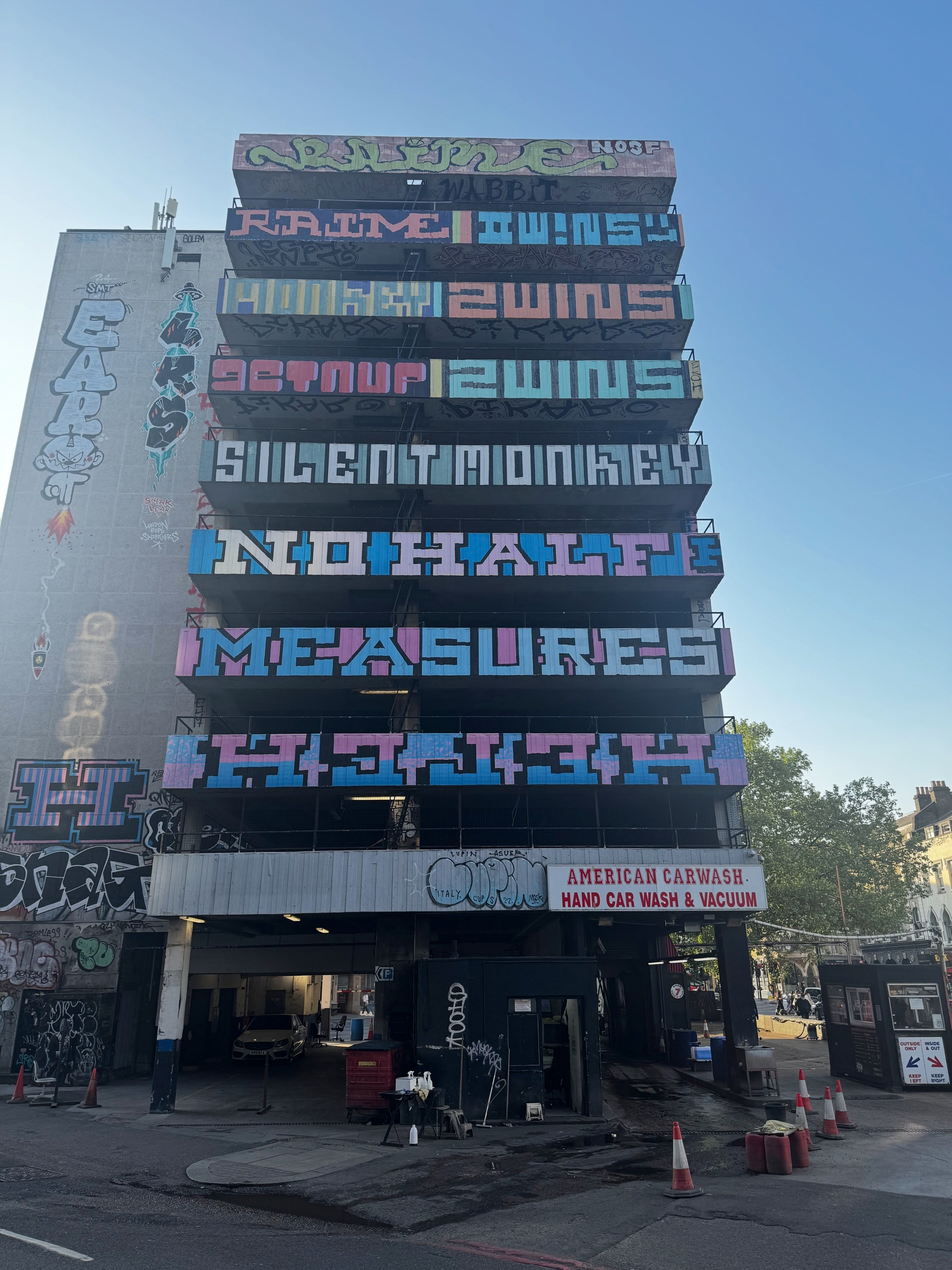 Multi-story concrete car park covered in large, colorful typographic art stacked across each level, with graffiti at street level and traffic cones marking the entrance.
