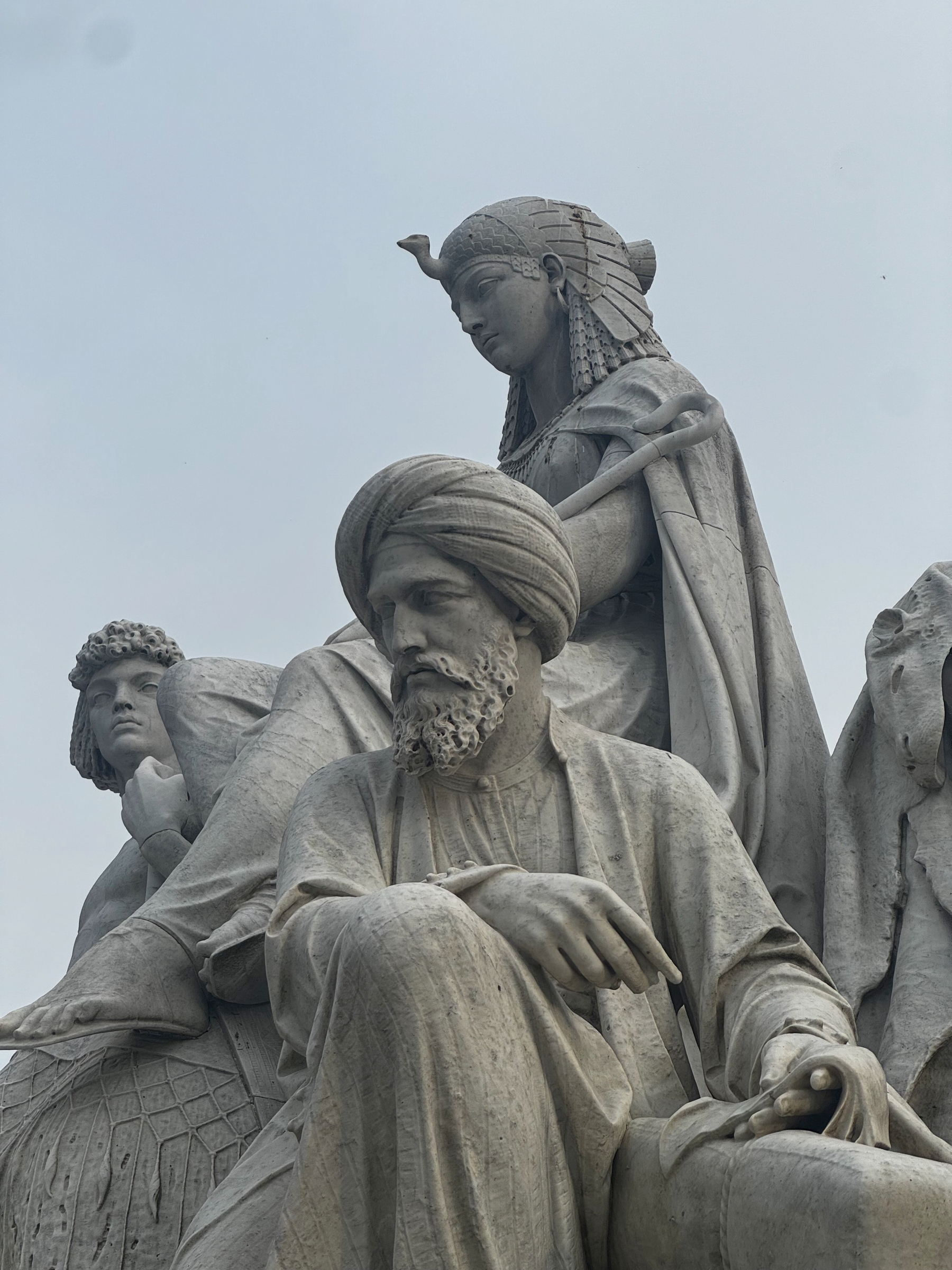 Close-up of pale stone statues with draped robes and carved faces, including a bearded figure seated in the foreground and others standing behind, their weathered surfaces and calm expressions giving a solemn, monumental feel against a grey sky.