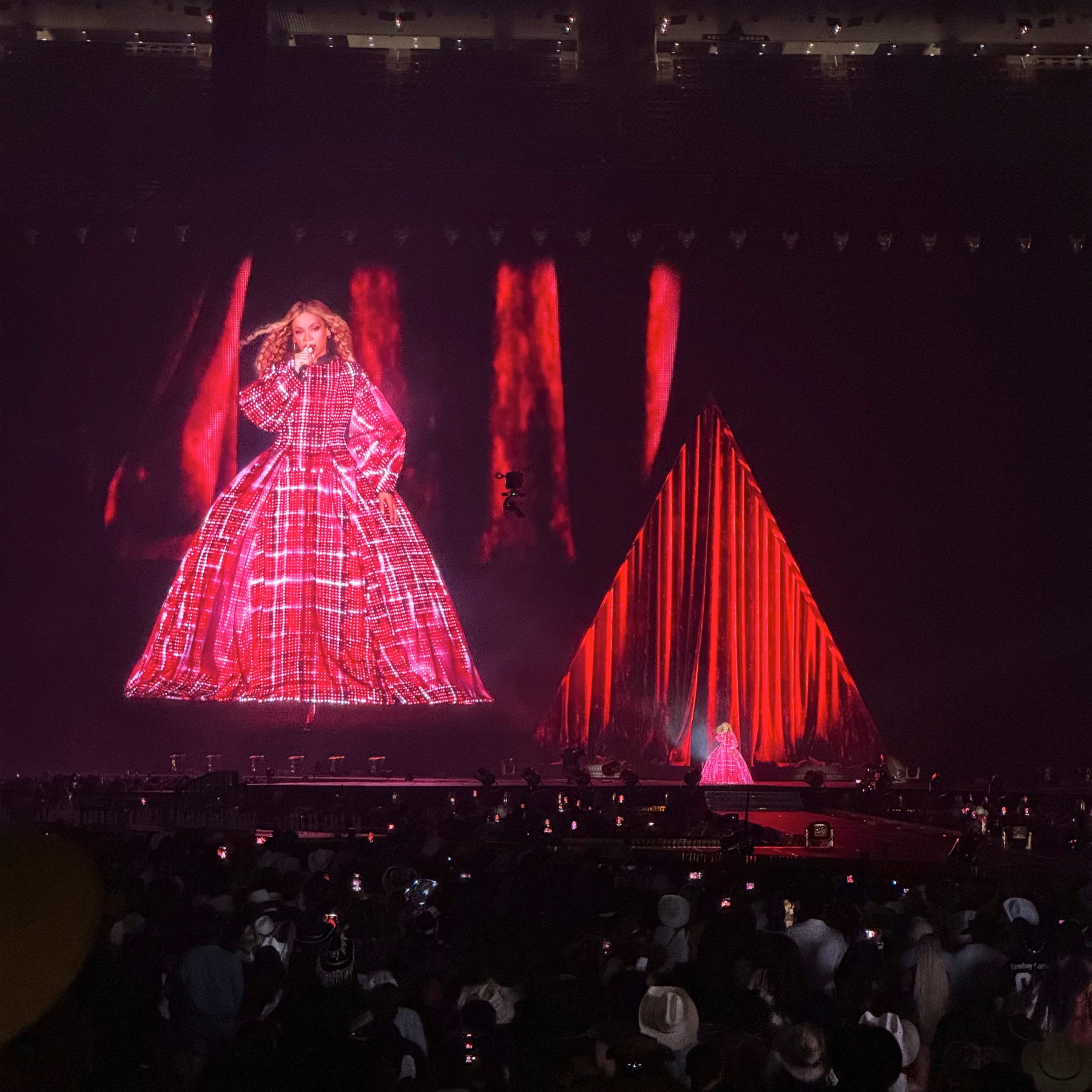 Beyoncé wears a striking voluminous red LED gown that fills the giant screen behind her as she sings, with a matching red triangular stage installation beside her. The rich crimson lighting and billowing fabric create a regal, cinematic moment against the packed stadium audience.