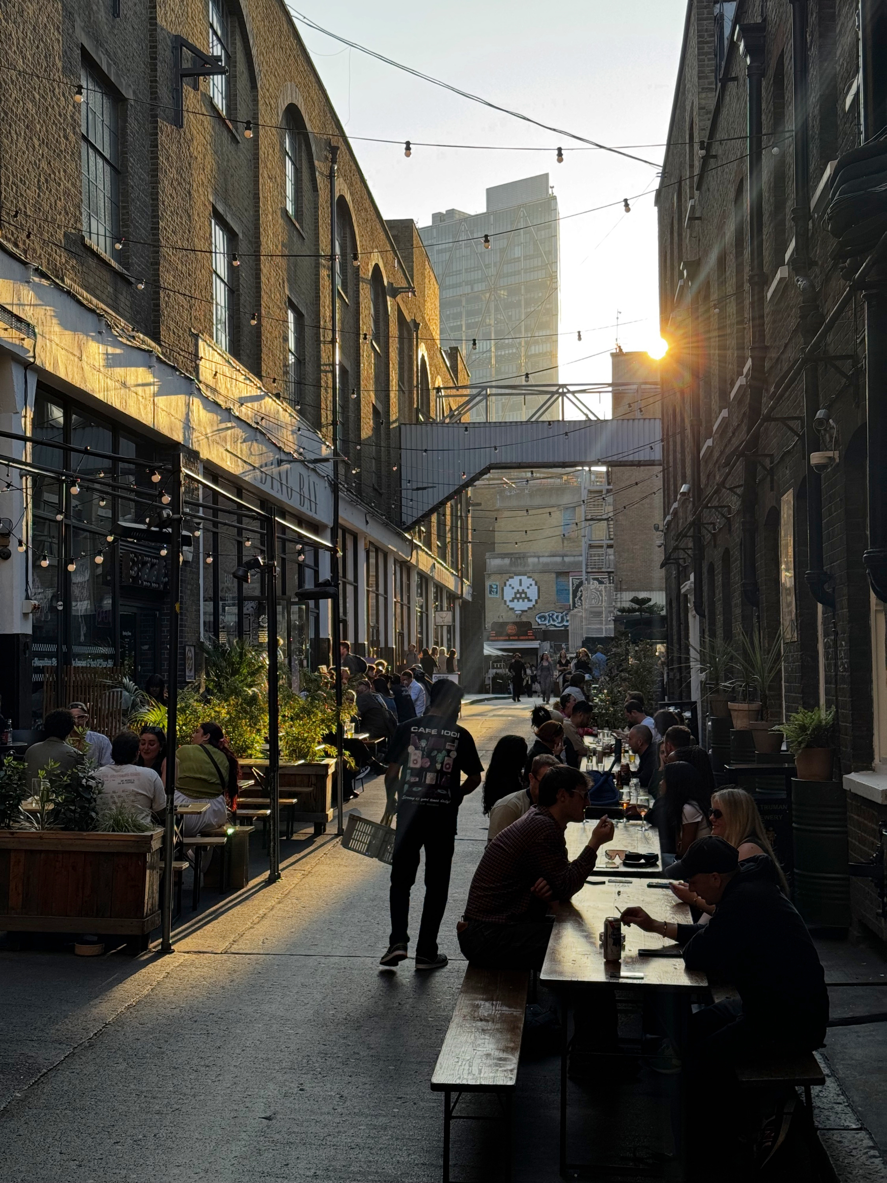 Crowded pedestrian lane lined with converted brick warehouses, long communal tables, hanging string lights, and people eating and drinking outdoors in warm late-afternoon sunlight.