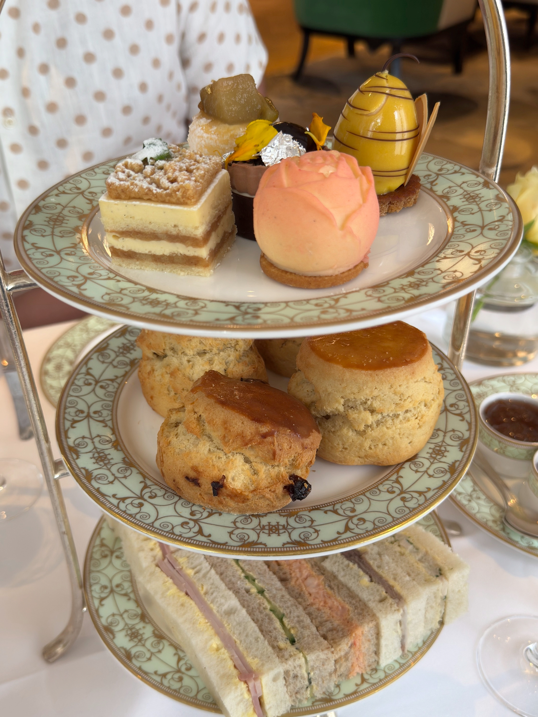 A three-tier stand arranged with neat finger sandwiches, golden scones with visible fruit, and an assortment of refined pastries. The layers feel abundant but orderly, with glossy finishes and precise shapes that invite lingering bites.