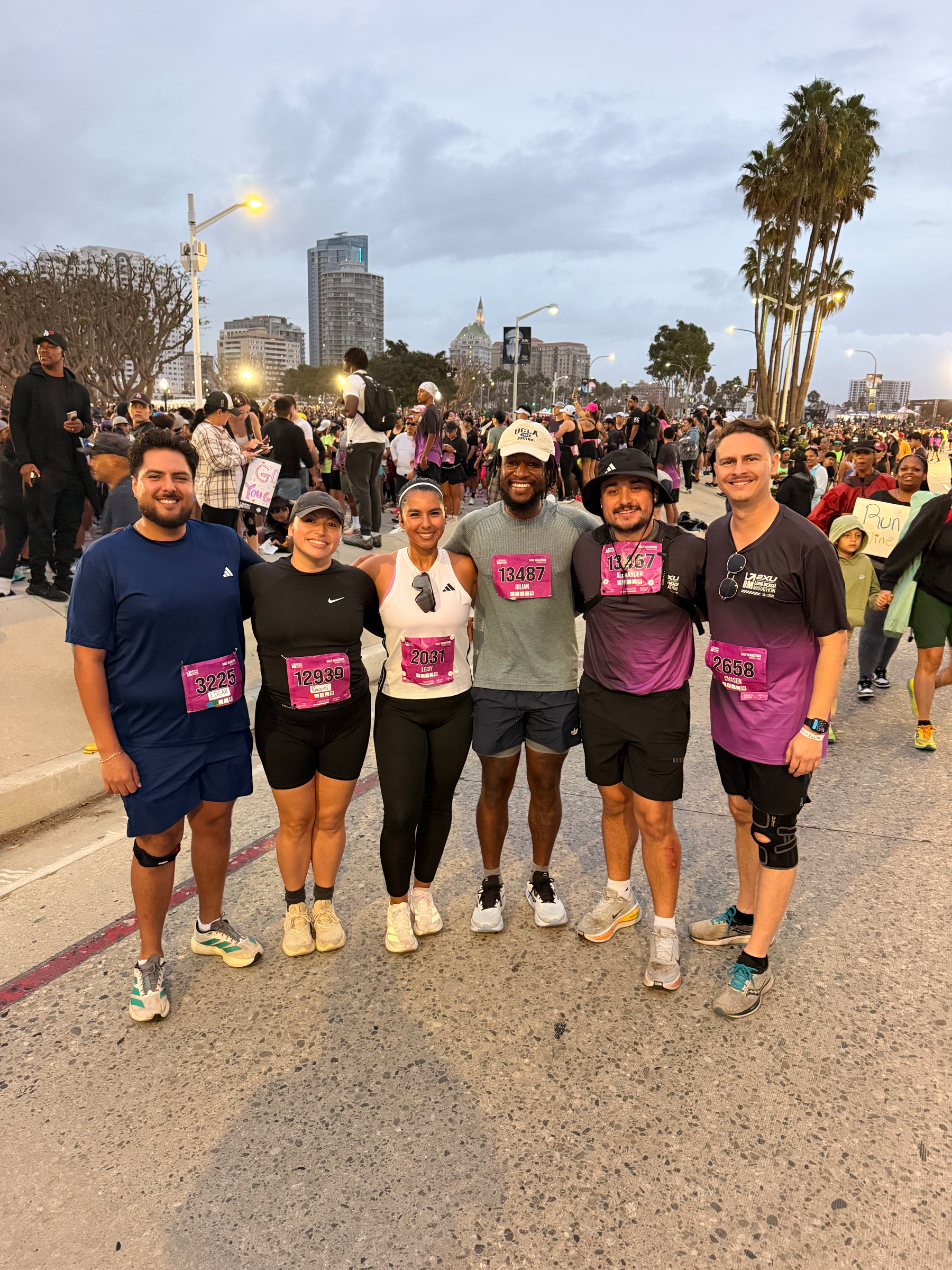 Our group of six runners wearing numbered race bibs and posing / smiling together on a city street during a large outdoor running event with many other participants in the background.