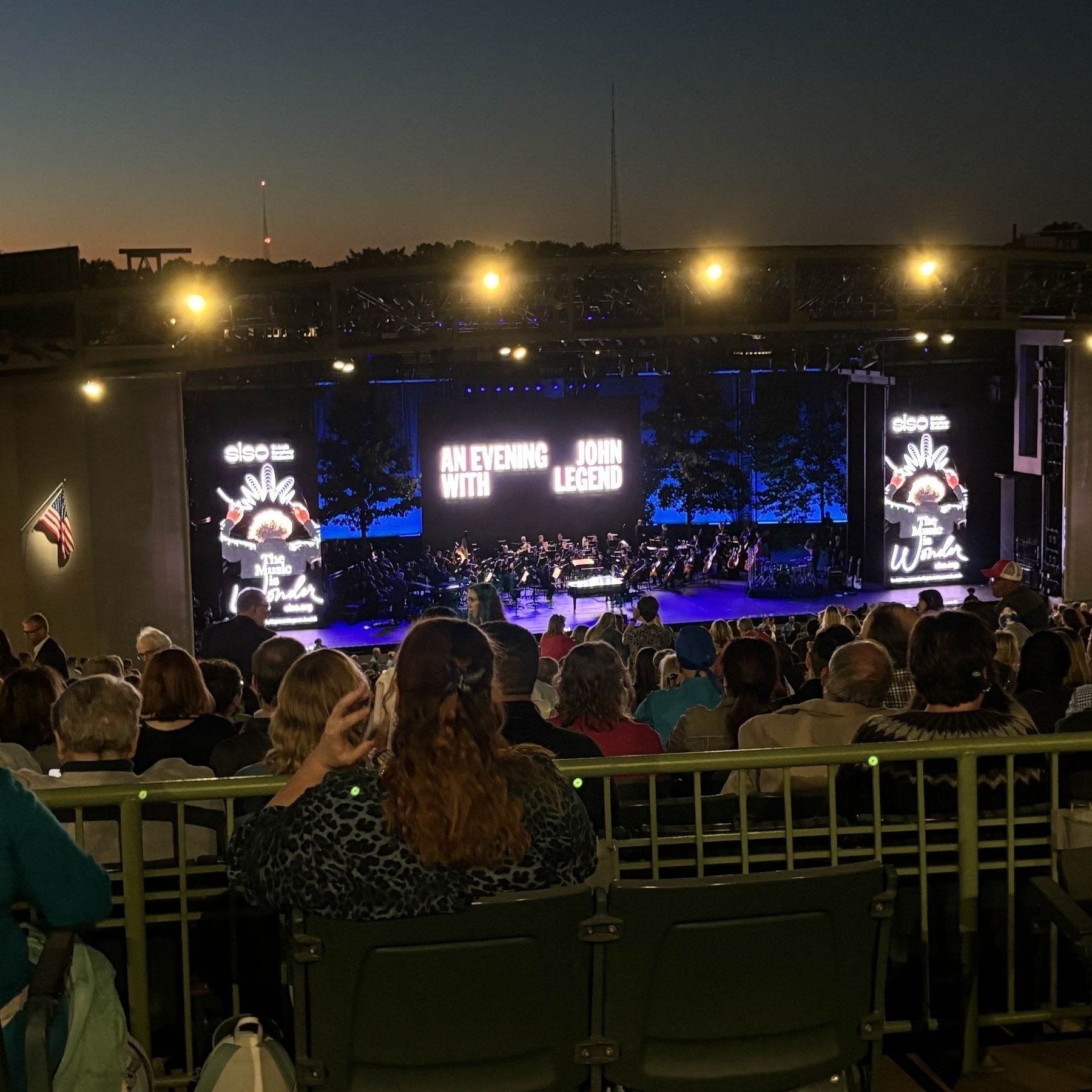 An outdoor stage, sunset in the background, with an orchestra assembling. A big sign in the middle reads: An Evening With John Legend