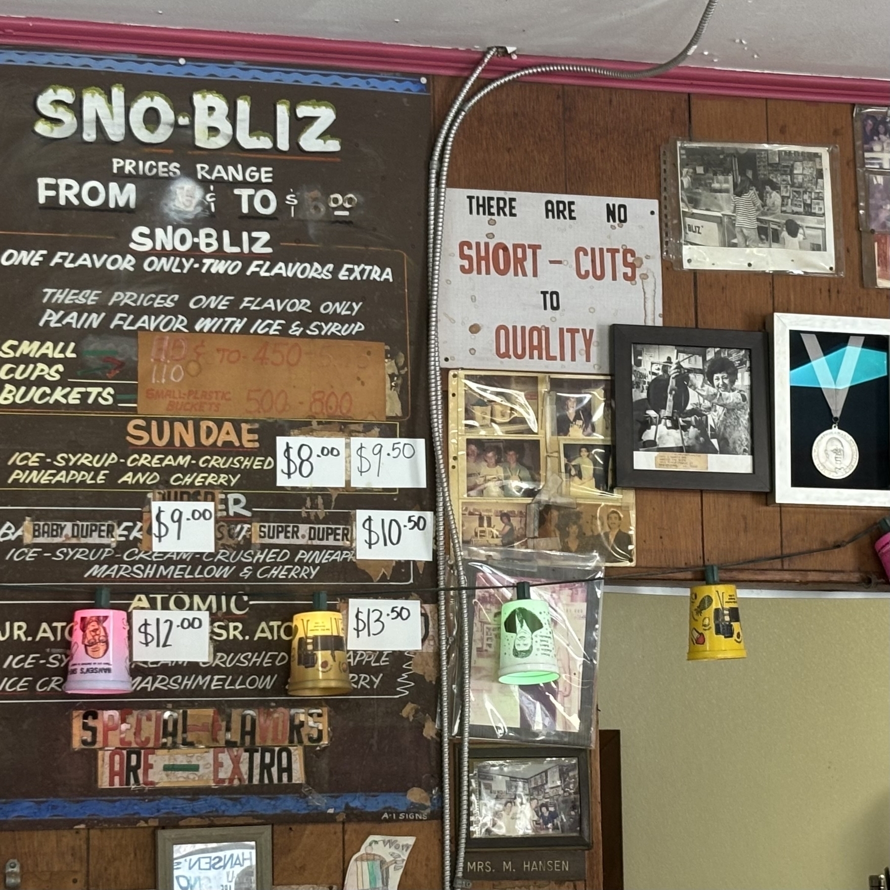 The wall of Hansen's Sno-Bliz on Tchopitoulas St. in New Orleans, LA. It is a mosaic of various signs and framed images, including: a menu, black & white photographs, a framed James Beard Award, and a sign that reads, 