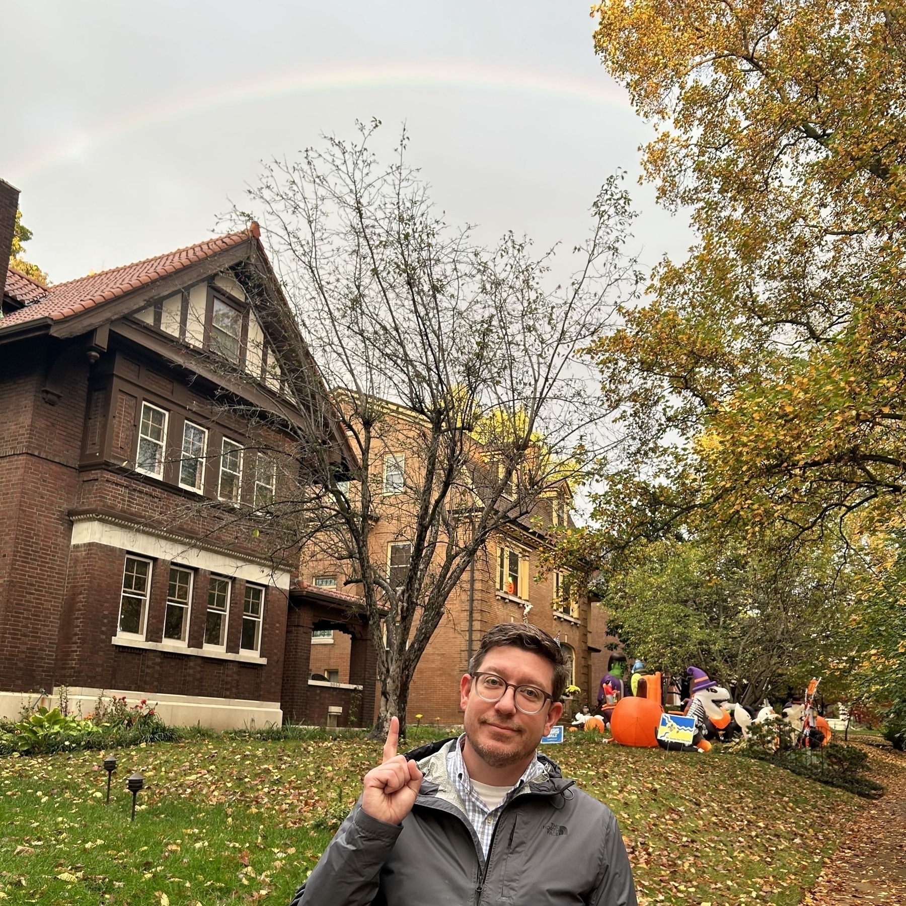 An average white male (me) wearing glasses and a gray raincoat pointing toward the sky with his index finger, pointing out a rainbow stretching over two houses in the background. 