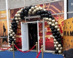 A balloon arch made of black and gold balloons decorates the entrance to an event, with red rope barriers leading to the doorway.