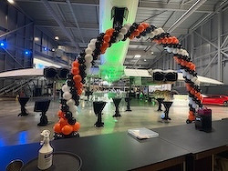 An aircraft in a hangar is decorated with an arch of black, white, and orange balloons, surrounded by tables set for an event.