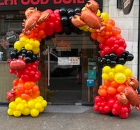 A festive archway made of red, orange, yellow, and black balloons, adorned with crab decorations, leads into a building.