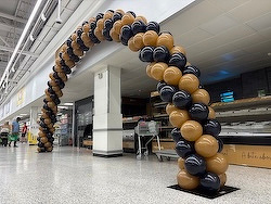 A balloon arch made of brown and black balloons is set up in an indoor space near shelves and a person walking by.