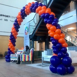 A large arch made of orange and blue balloons decorates an indoor space, with a person seated below and a staircase in the background.