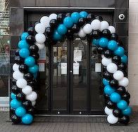 A balloon arch made of black, white, and teal balloons decorates a glass door entrance.