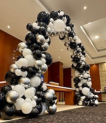 A large arch made of black, white, and silver balloons is set up in a room with a high ceiling.