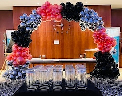 A decorative arch made of pink, black, and silver balloons stands behind a table filled with empty glasses, set against a wooden backdrop with a clock on the wall.