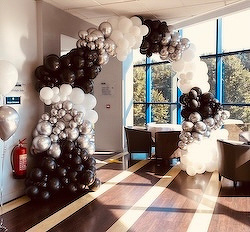 A balloon arch made of black, white, and silver balloons decorates a room with a table and chairs, next to large windows.