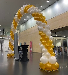 A balloon arch made of gold and white balloons stands in a modern indoor space with high ceilings and sleek decor.