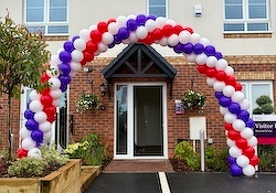 A colorful balloon arch in red, white, and purple decorates the entrance of a building surrounded by plants and a driveway.