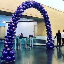 A balloon arch in shades of blue and purple stands in a modern indoor space with people walking nearby.