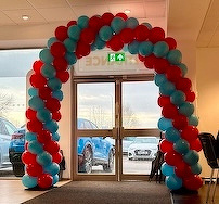 A colorful balloon arch of red and blue balloons decorates the entrance of a room with glass doors, overlooking parked cars outside.
