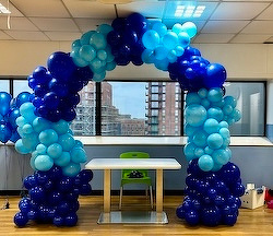 A balloon arch made of various shades of blue balloons is set up in a room with a small table and a green chair, overlooking a cityscape.