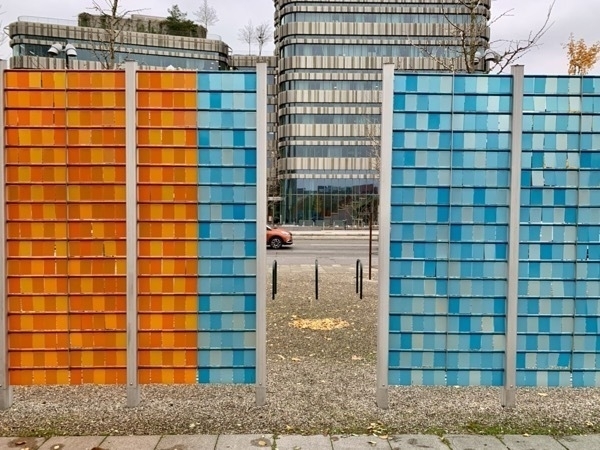 Artistic orange and blue divider with Malmö University in the background and visible through a gap in the fence