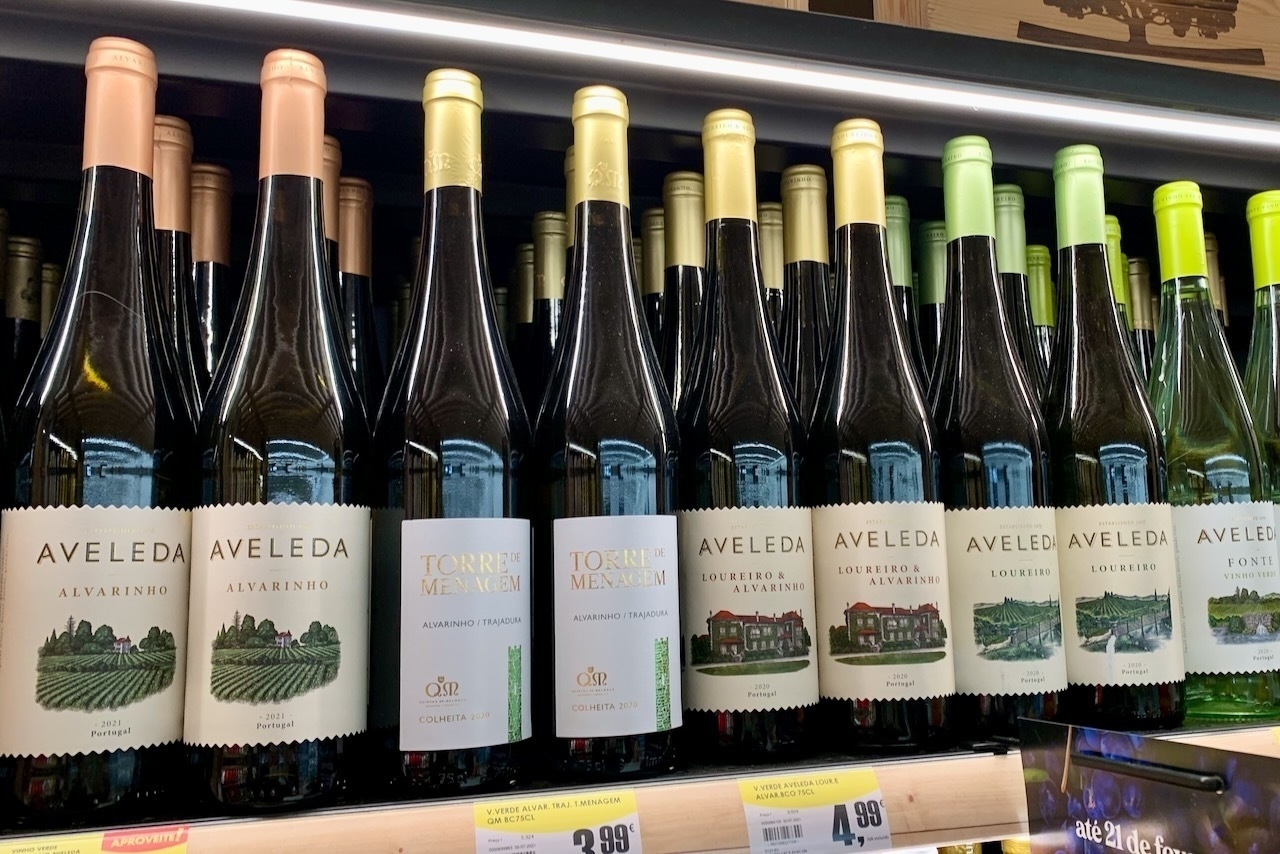 A row of wine bottles, mostly of a Portuguese label ‘Aveleda’ with colourful metallic foil capsules in rose gold, yellow and green.