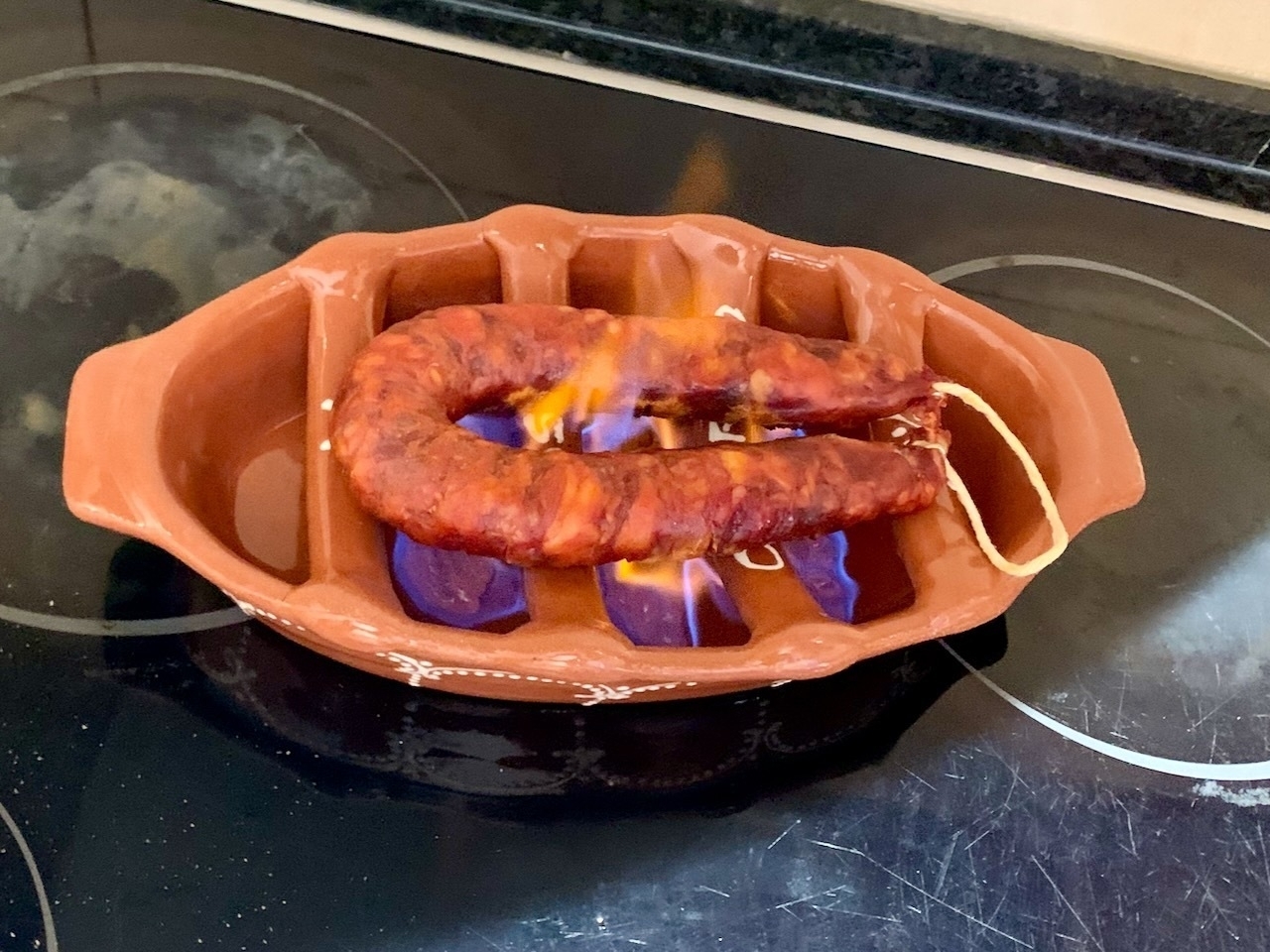 An ‘assador’, a Portuguese ceramic dish with bars across it, filled with lighter fluid gel that’s been set alight, grilling a chouriço Portuguese sausage.