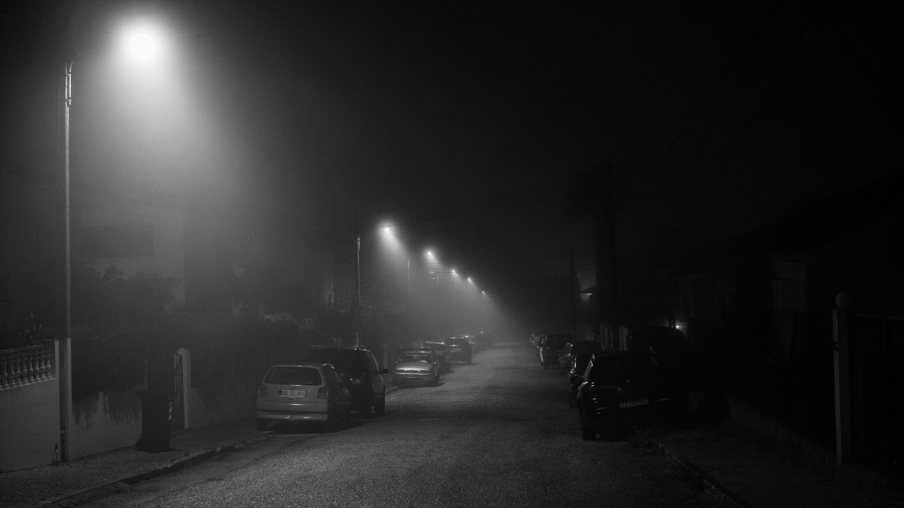 Looking down a residential street, several cars parked on the side of the road. The light from a row of street lamps are diffused through fog.