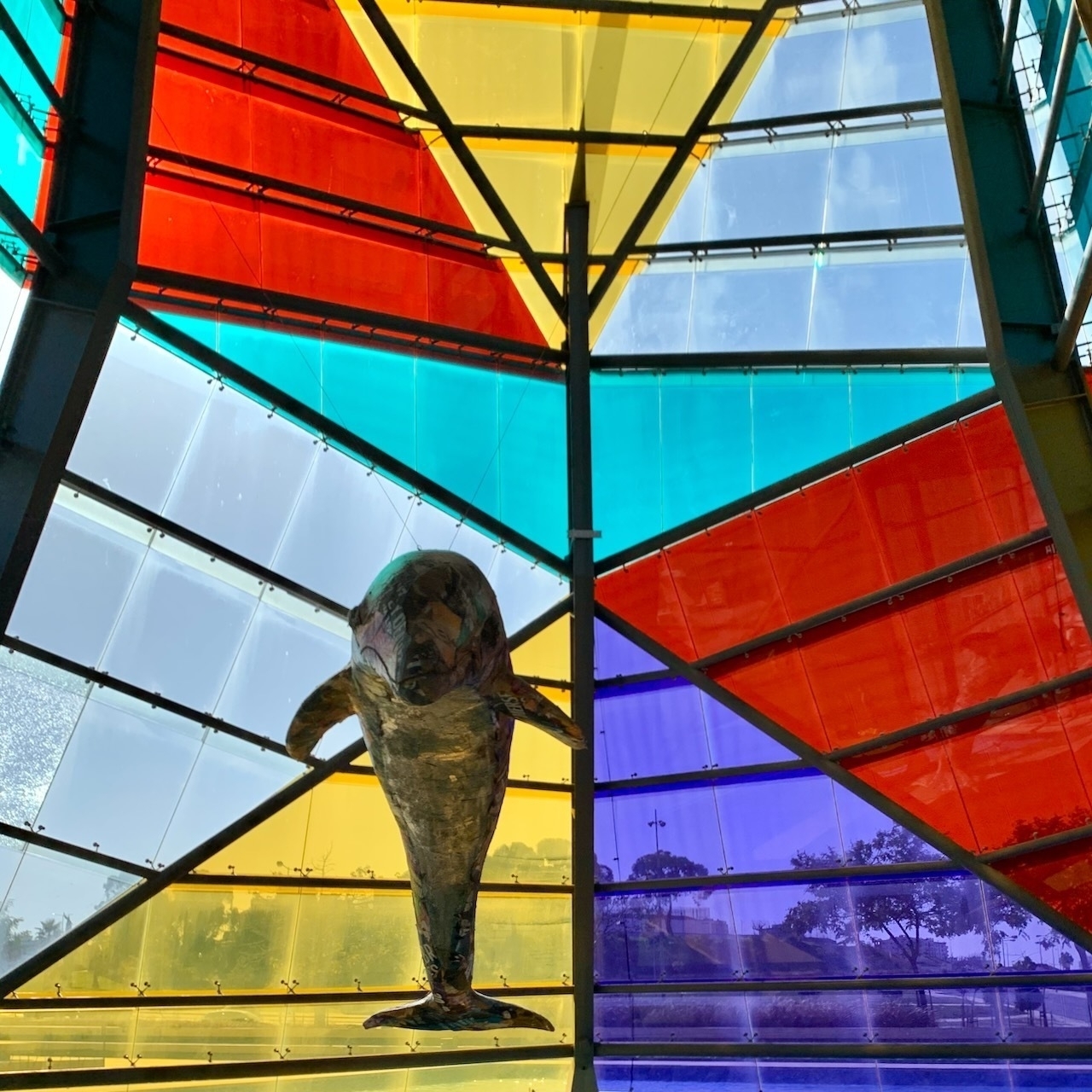 Modern geometric stained glass panels over an entry foyer at a shopping mall in Setúbal, Portugal. There is a dolphin sculpture suspended in the foreground