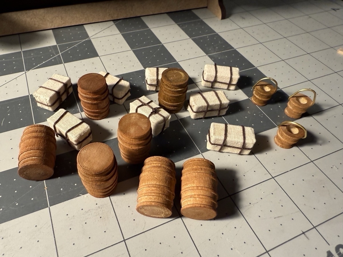 A pile of cargo to be stored in the lower hold of the flying dutchman model ship. Shown are several wooden barrels, a few wooden buckets with brass handles, and several packages wrapped in sail cloth and tied up with brown rope. 