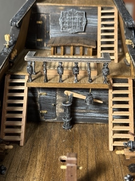 View of the flying dutchman model ship looking toward the stern. In it you see three ladders. Two going up to one deck and another going up one level higher. Also shown are a bilge pump, bell and an additional rail installed as part of this step. 