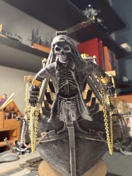 The skeletal figure head of the flying dutchman model ship mounted to the prow. You can see the brass manacles around each wrist with chains hanging down and mounted to the hull farther back. 