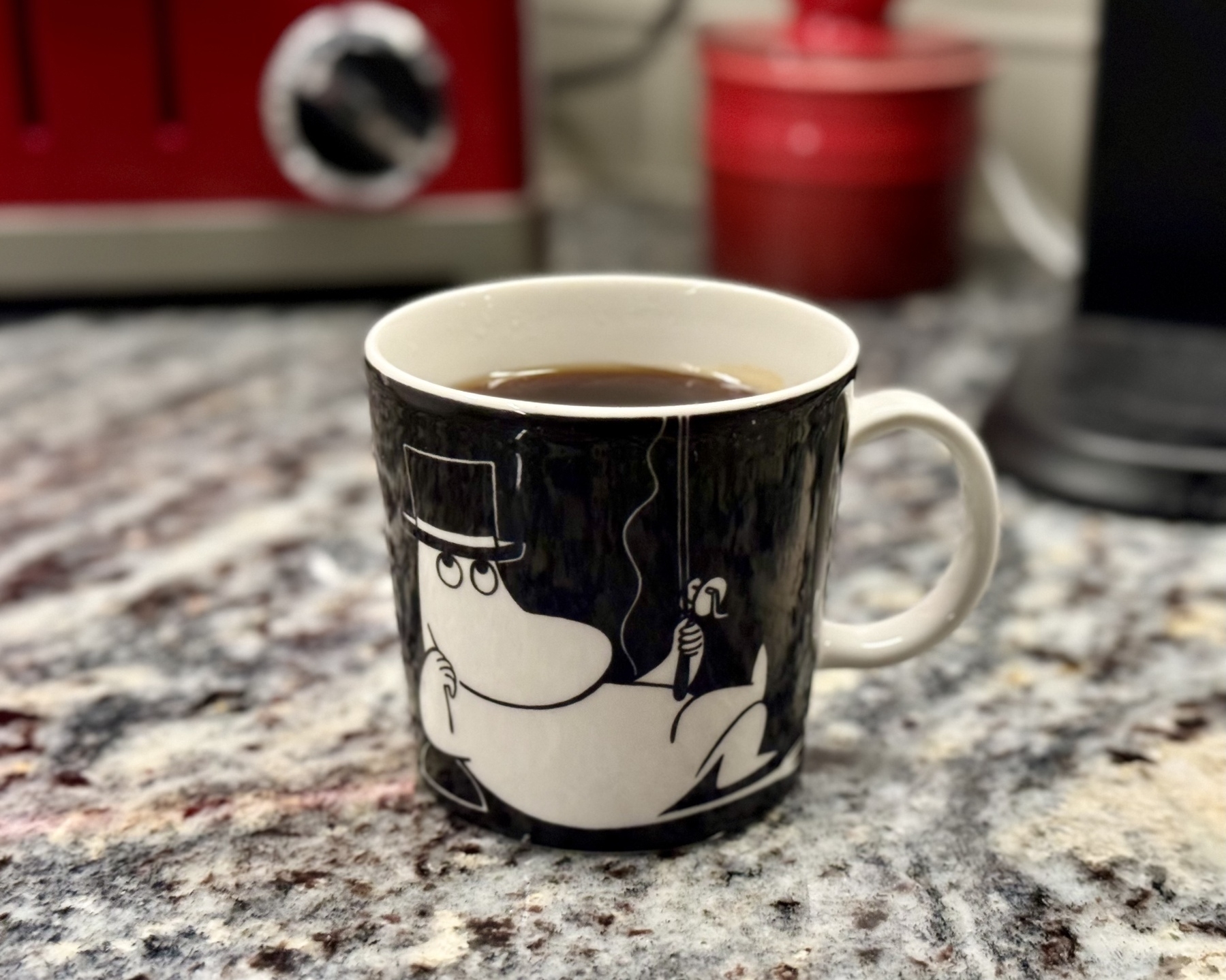 A cup of coffee on a kitchen countertop. Drawn on the mug is Moominpappa, a character from Finnish children’s books.
