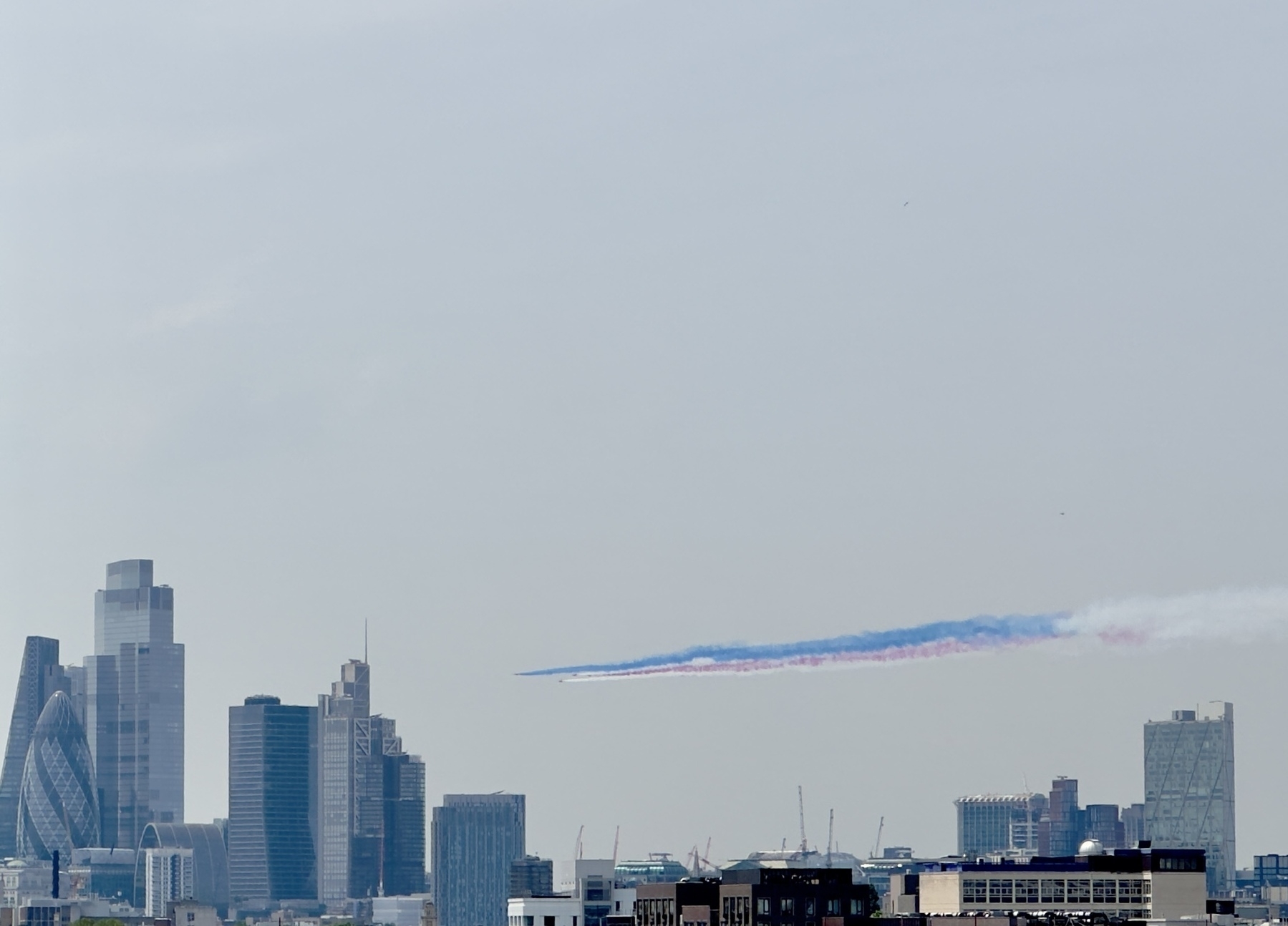 Jets from the Red Arrows flying over the City of London, trailing red, white, and blue smoke.