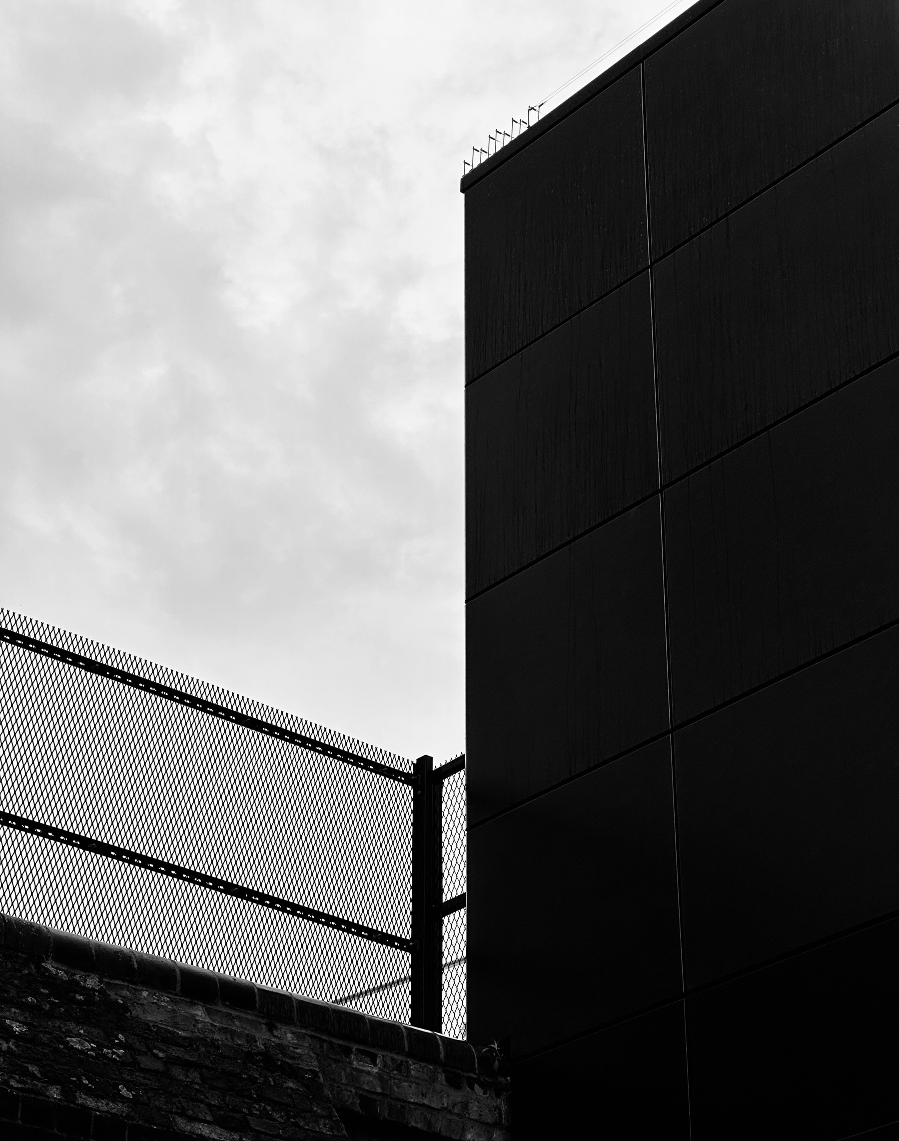 Two walls meeting at a right angle, one made of brick and one made of black panels. A grey sky above.