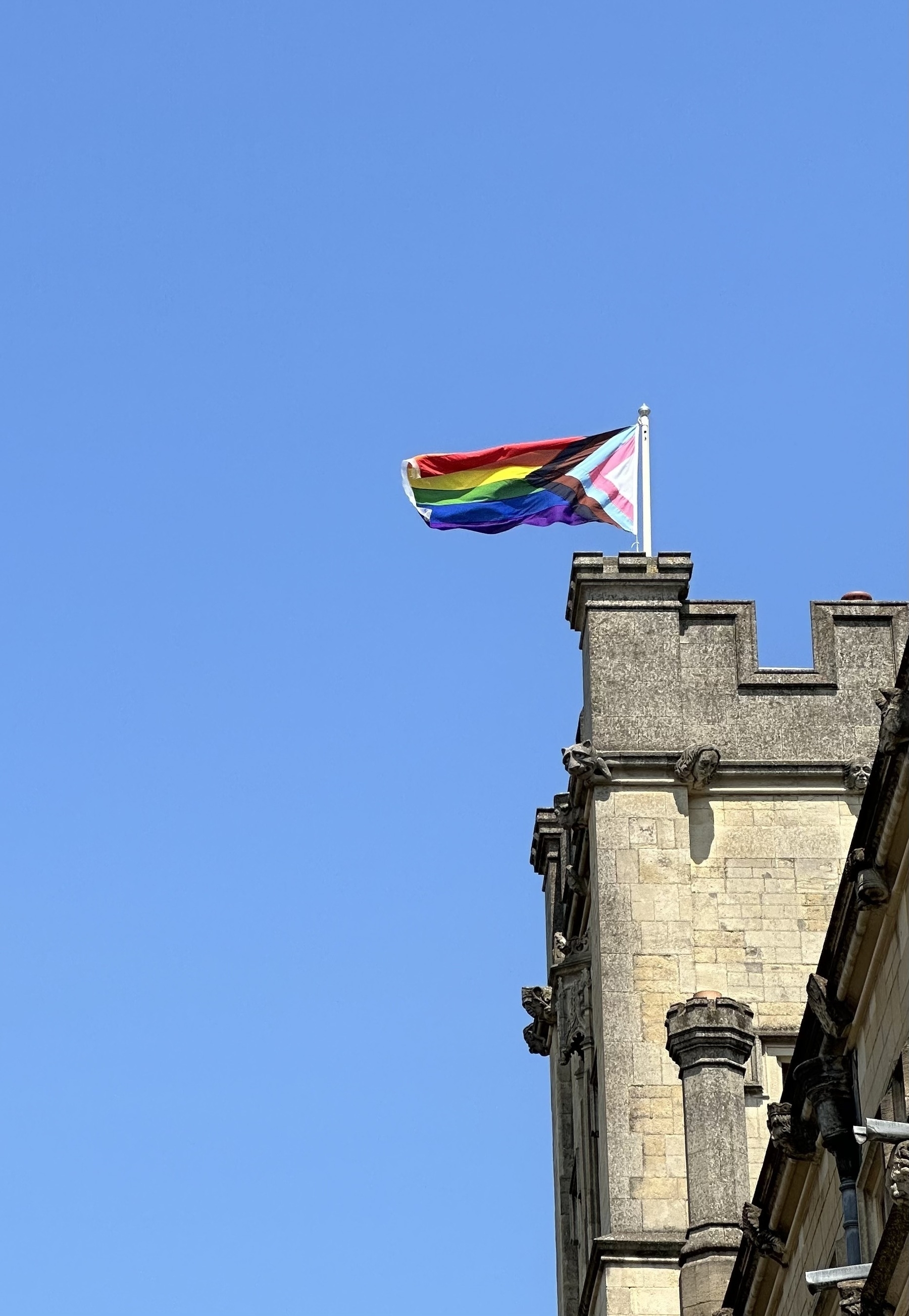 A progress pride flag flying from the top of a stone tower in Oxford, against a clear blue sky.
