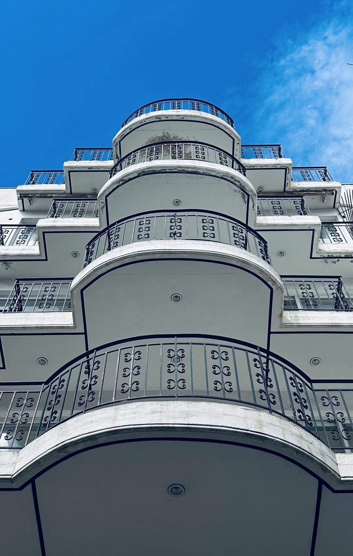 Looking up at a white and black art deco building with a series of balconies, underneath a blue sky.