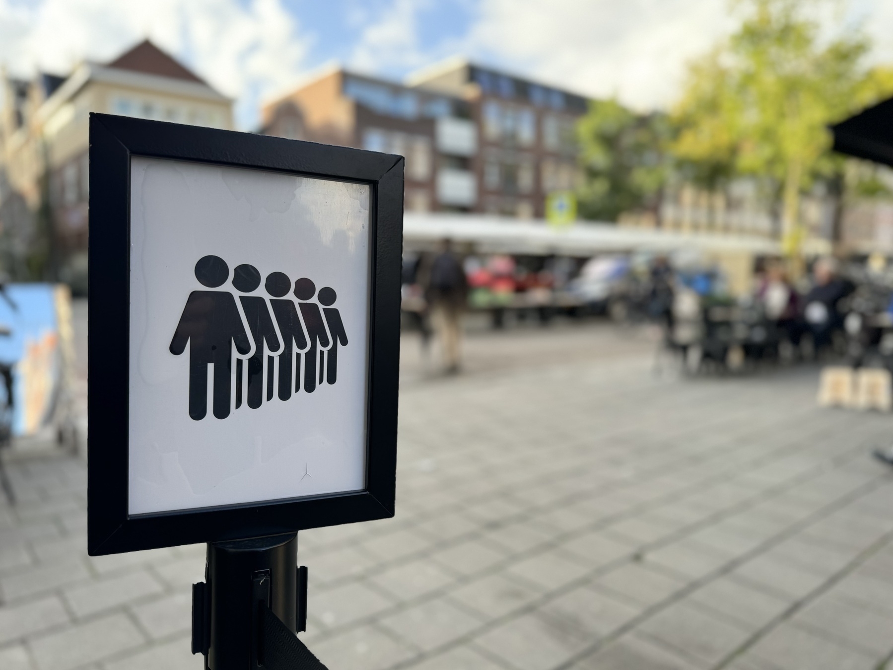 A sign showing people queueing with an out of focus background of an outdoor market.