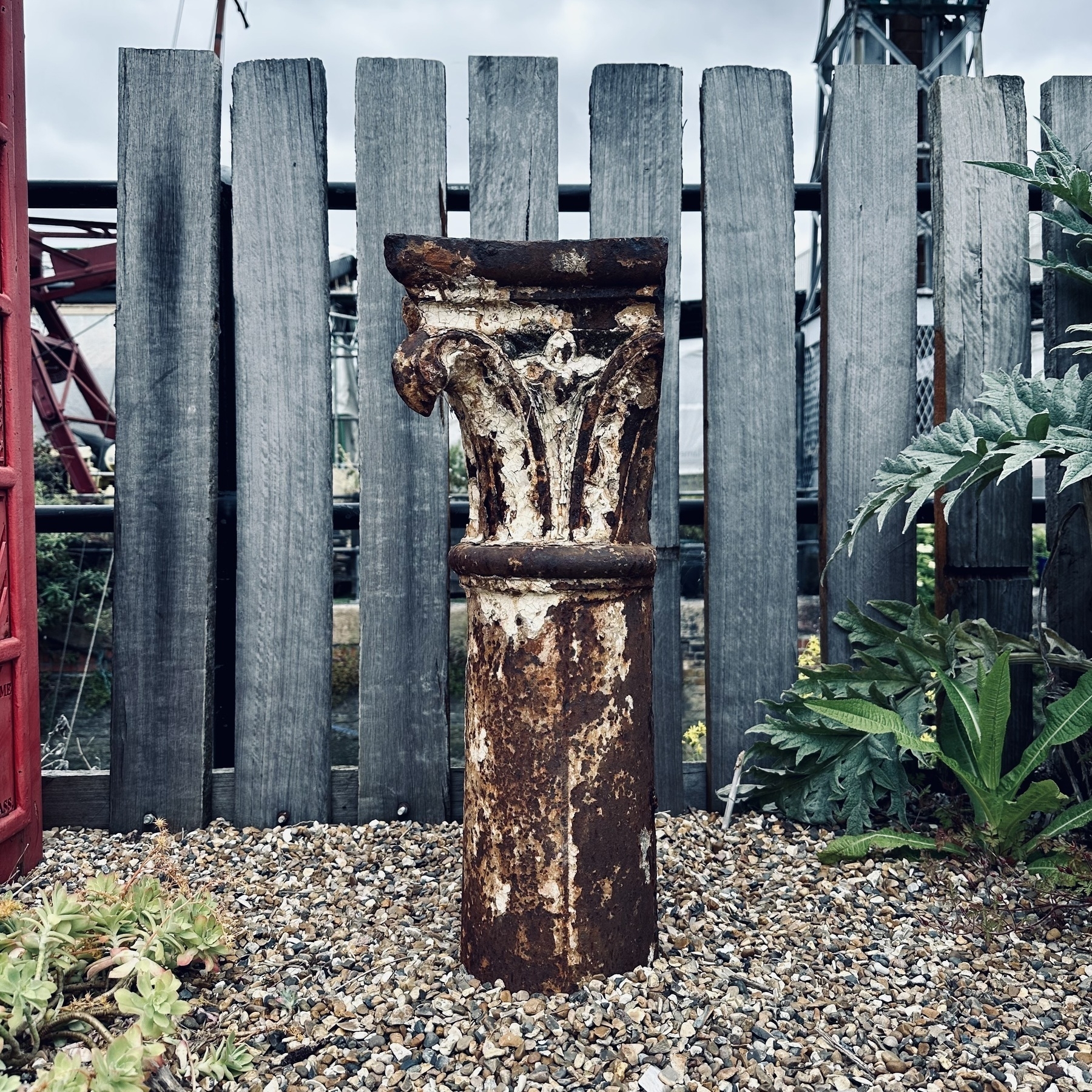 The top of a rusted metal Corinthian column, embedded in gravel, with a wooden fence behind.